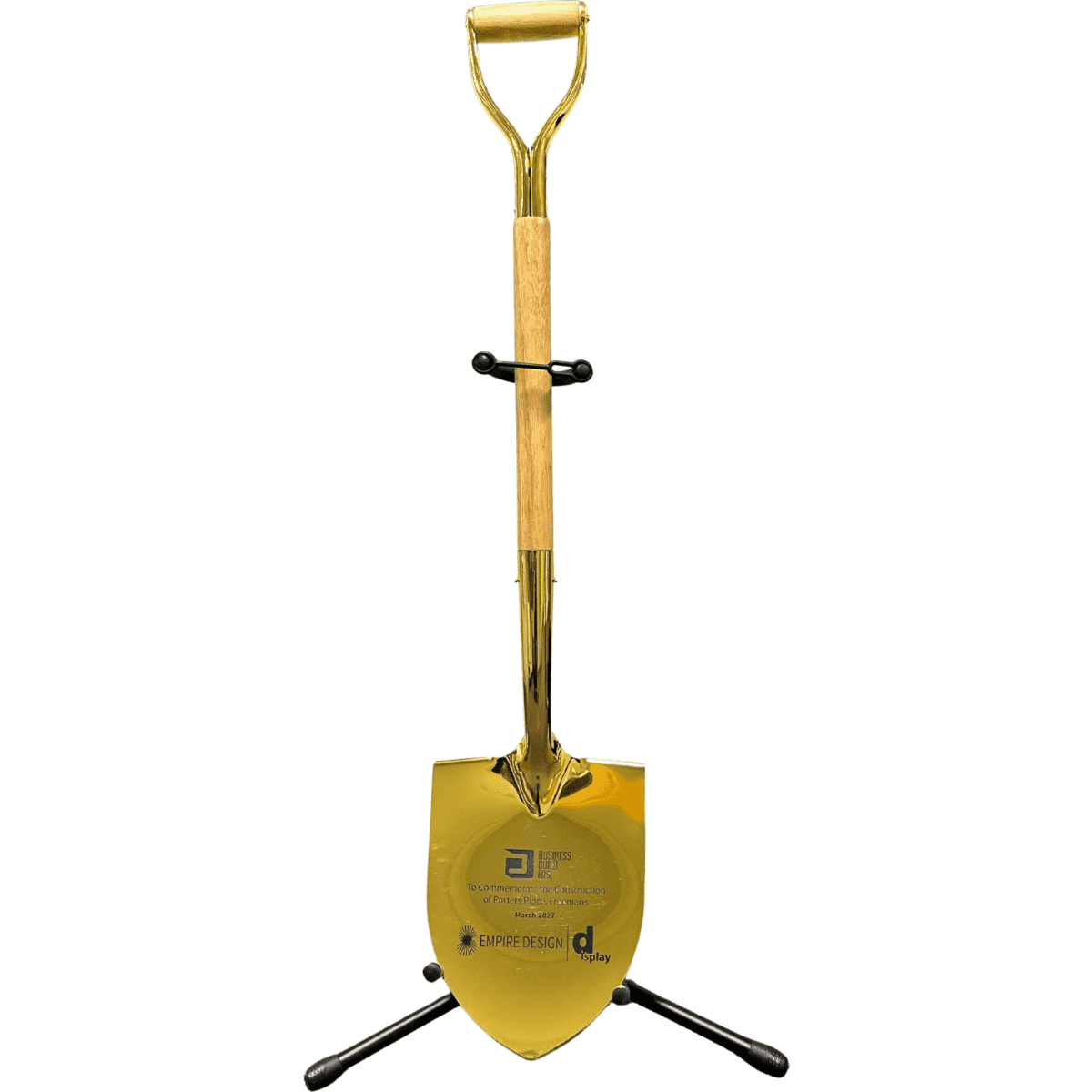 A shiny gold shovel with a wooden handle, displayed upright on a black stand. The stand has a logo.