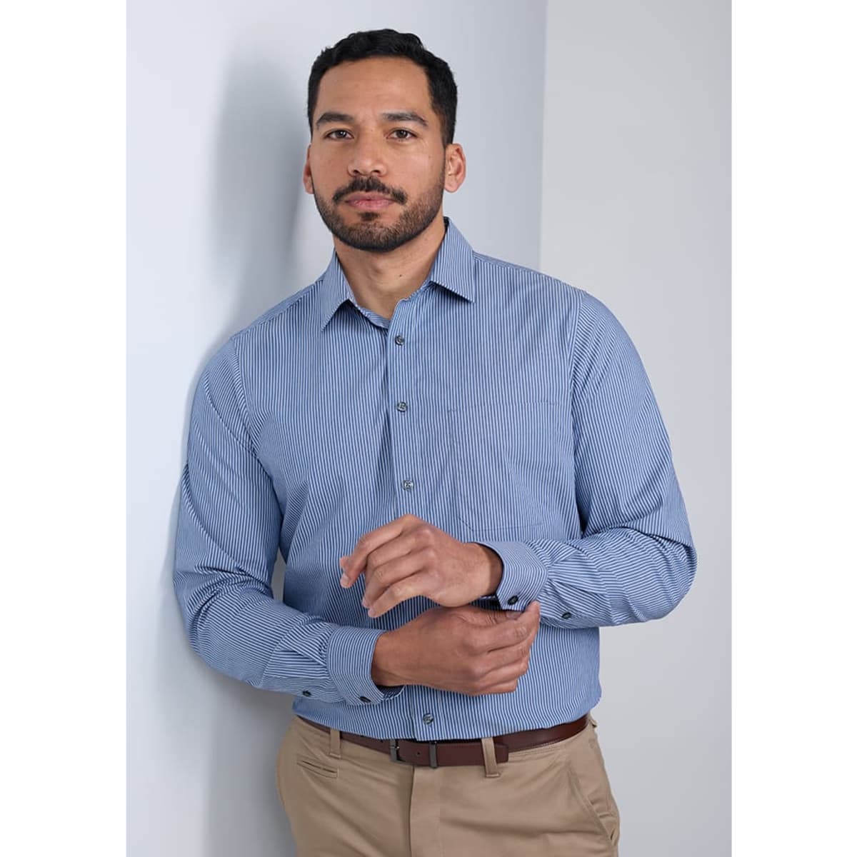 A man stands against a wall wearing a blue and white striped long sleeve shirt, paired with beige trousers.