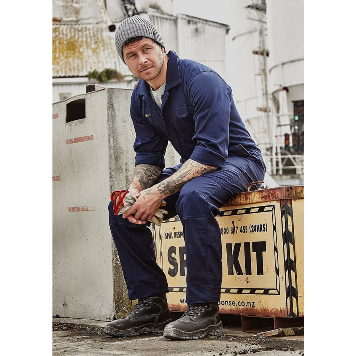 A man in a navy blue lightweight cotton drill overall, seated on a box outside, wearing a beanie and work boots.