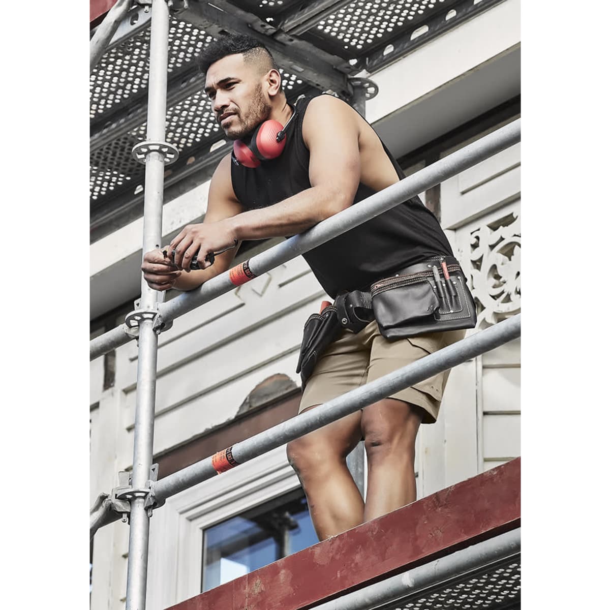 A man in a black sleeveless tee and shorts stands on scaffolding, wearing headphones and a tool belt.
