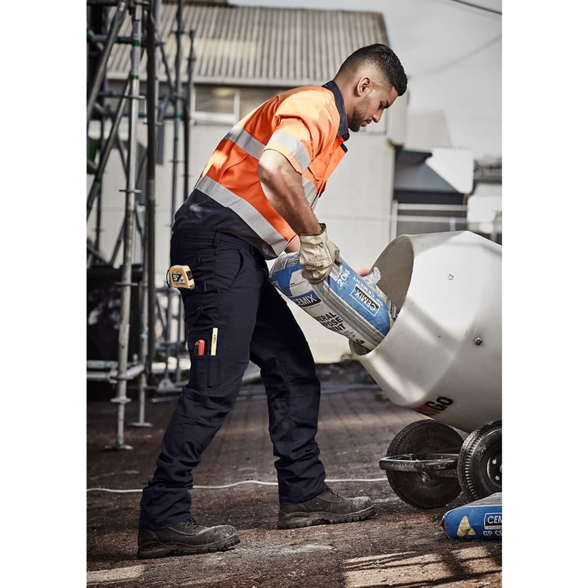 A man in orange and grey high-visibility workwear pours from a bag into a mixer, wearing navy rugged cargo pants.