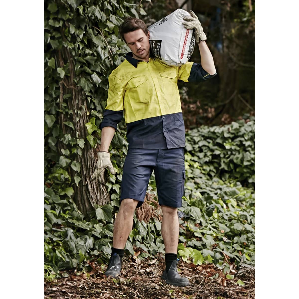 Men's rugged cooling vented shorts in navy, styled with a bright yellow shirt, set against a leafy background.