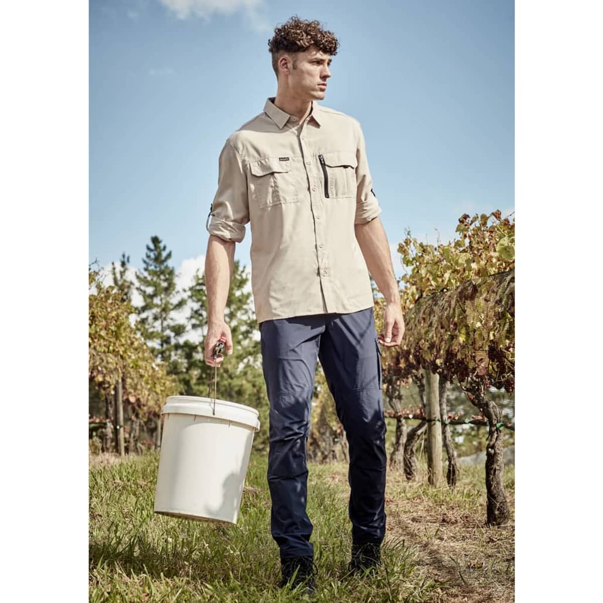 A long-sleeve, button-up shirt in beige, worn with navy pants, against a vineyard backdrop. Features a logo.