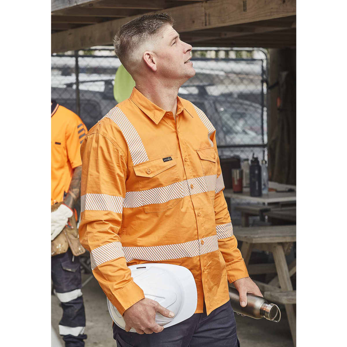 An orange long-sleeve work shirt with segmented reflective tape, featuring a pocket and a branding logo.