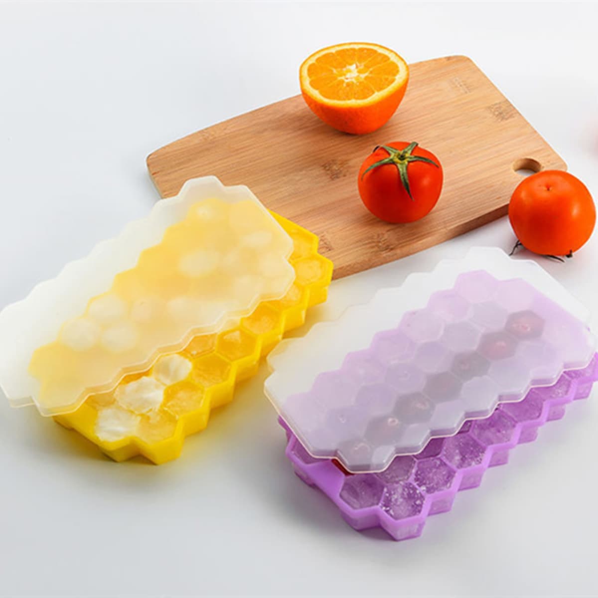 Hexagonal silicone ice cube trays in yellow and purple, with clear lids, on a wooden cutting board alongside fruits.