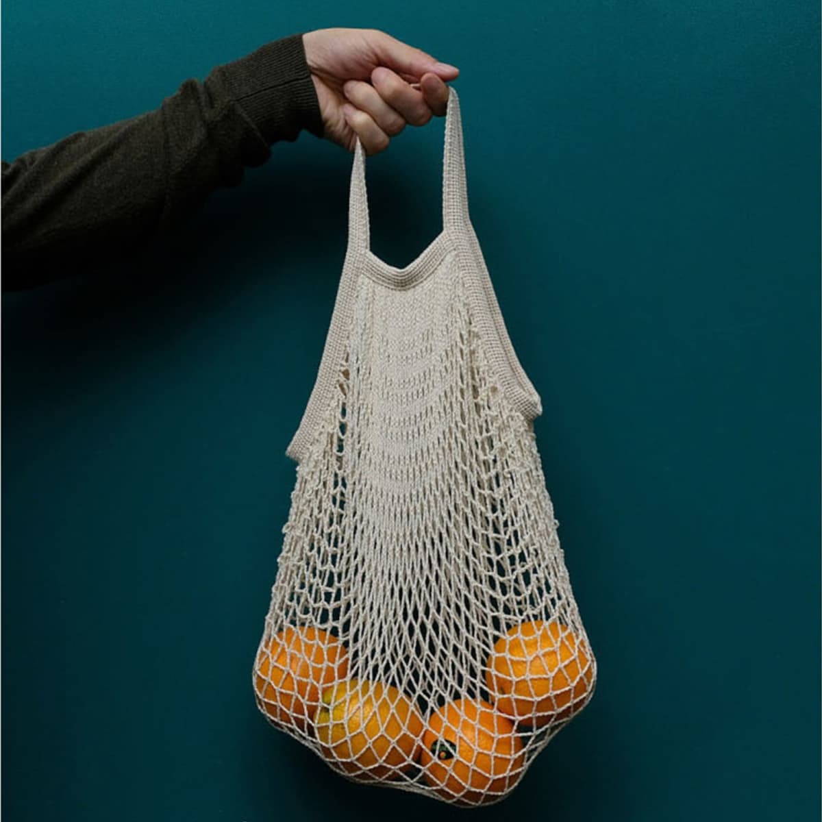 A hand holds a beige cotton mesh bag filled with oranges, set against a dark green background.
