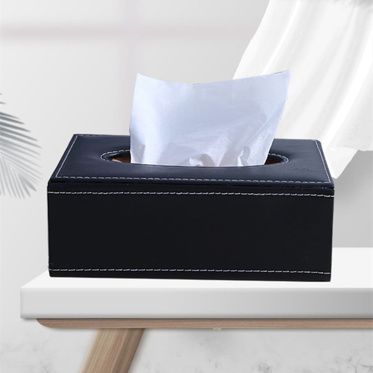 A black leather tissue box with white stitching, featuring a slot for tissues, placed on a light-coloured table.
