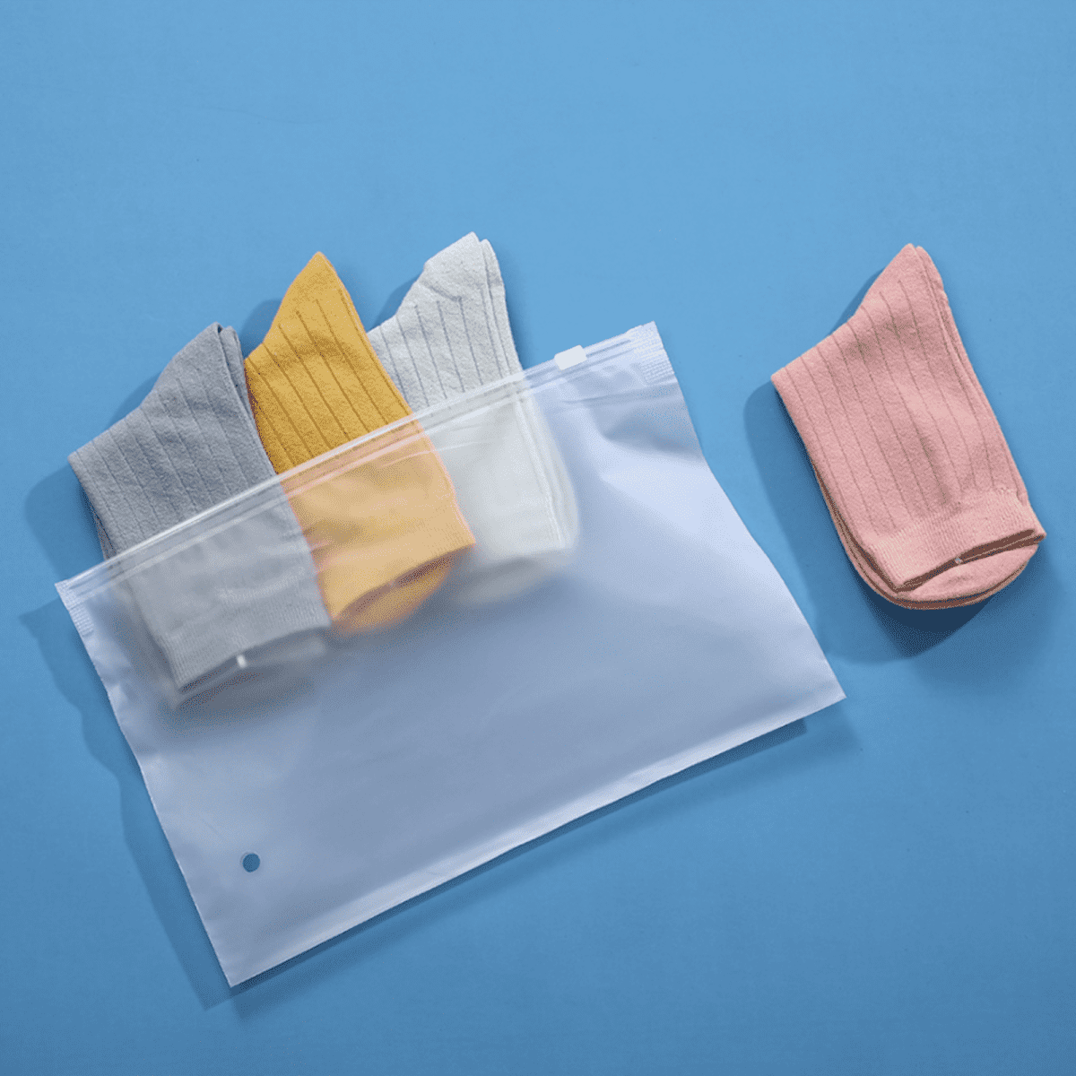 A matte frosted zip-lock storage bag containing grey, yellow, and white socks, with a pink sock placed beside it.