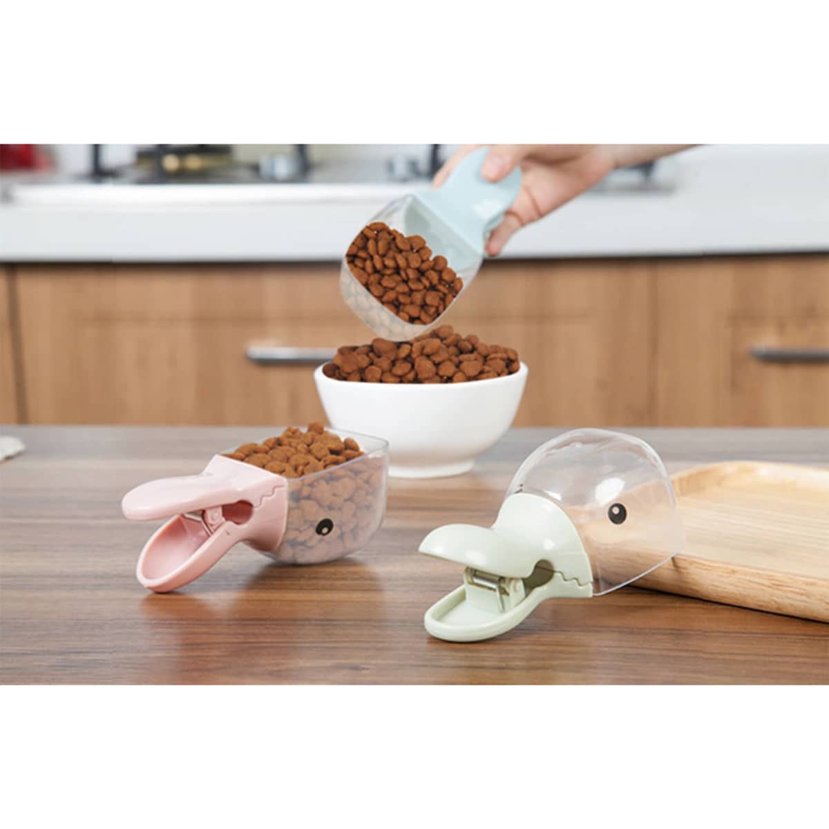 A pet food scoop in pink and green, with a clip, above a bowl of kibble on a wooden surface.