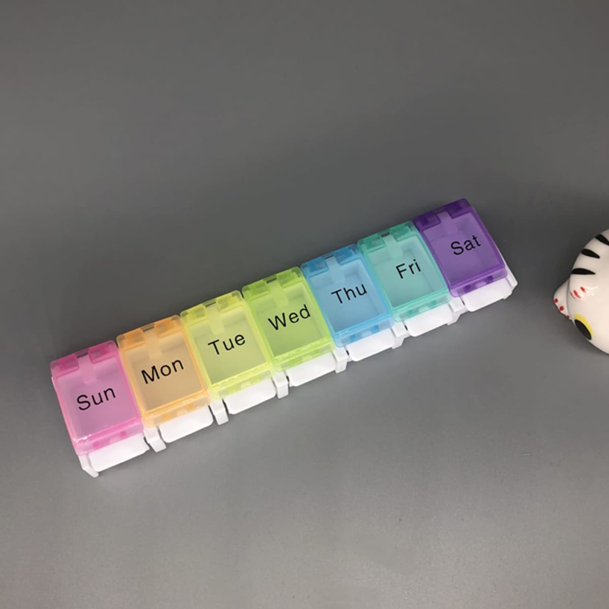 A multi-coloured seven-grid pill box featuring compartments for each day of the week. It has a white base and a logo.