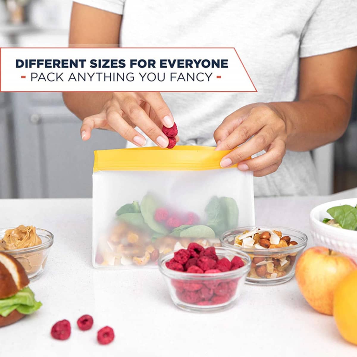 A food storage bag with a yellow seal is filled with nuts and raspberries, surrounded by bowls of fruit and snacks.