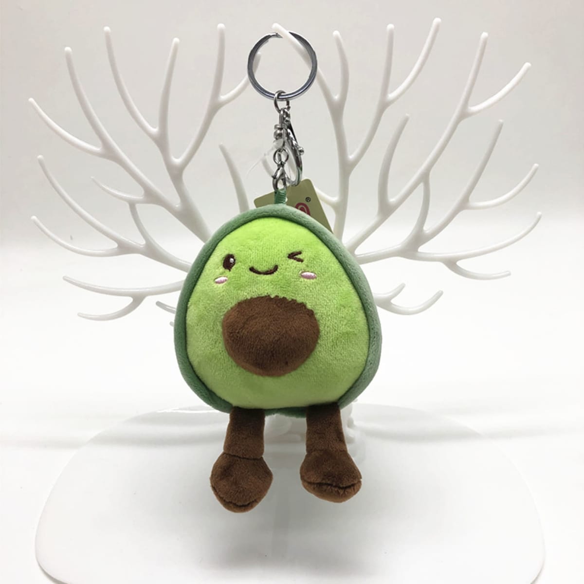 A green avocado plush key chain with a smiling face, hanging from a silver key ring against a white display stand.