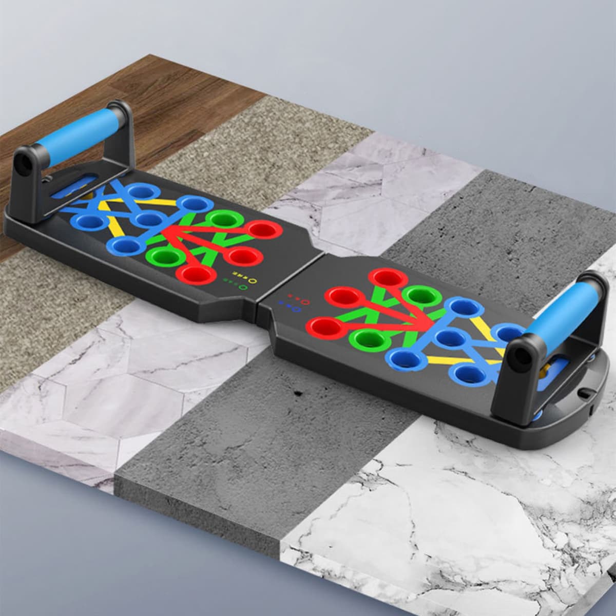 A push-up support board in black with blue handles and multi-coloured holes on a textured surface.