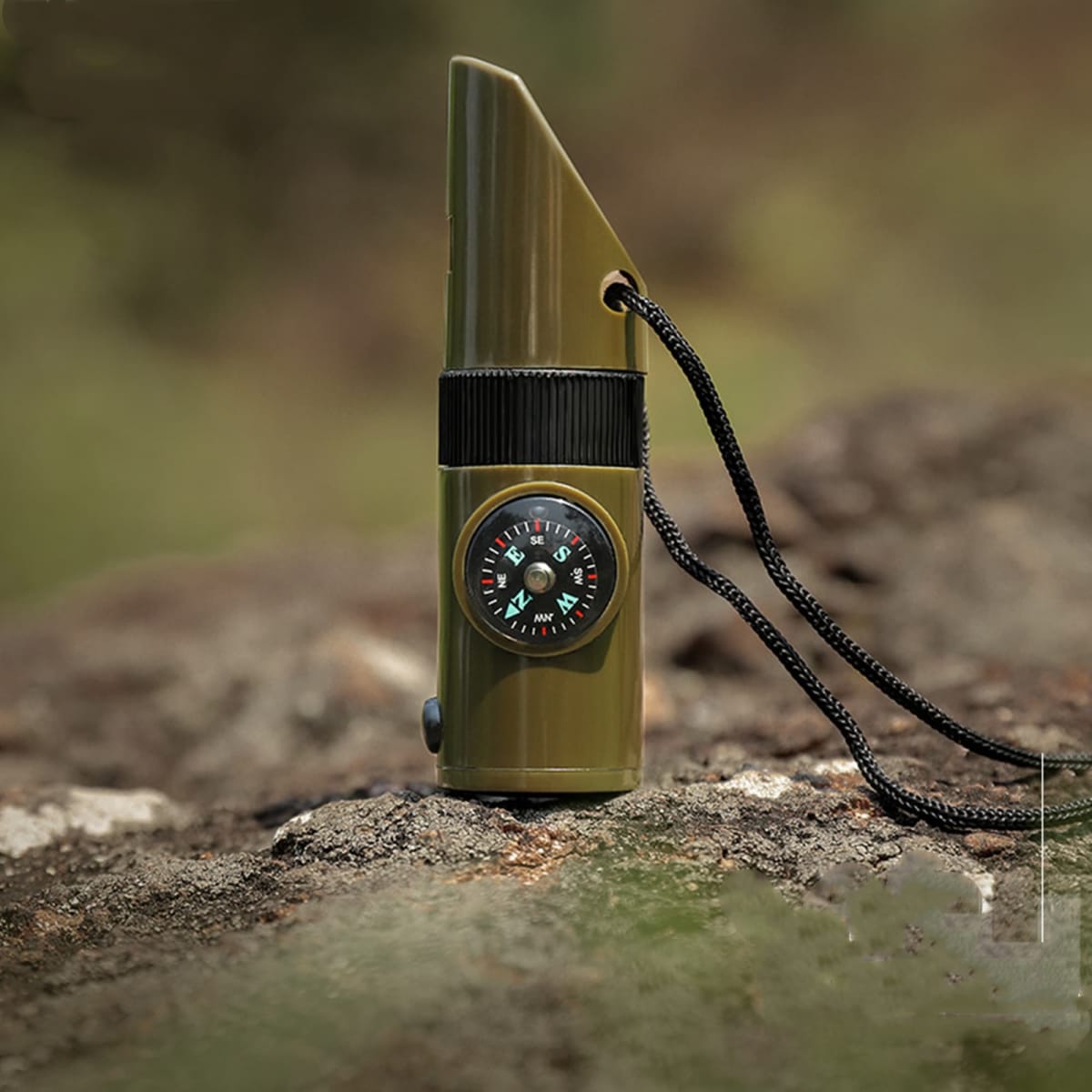 A green camping whistle with a built-in compass, attached to a black lanyard, resting on a rocky surface.