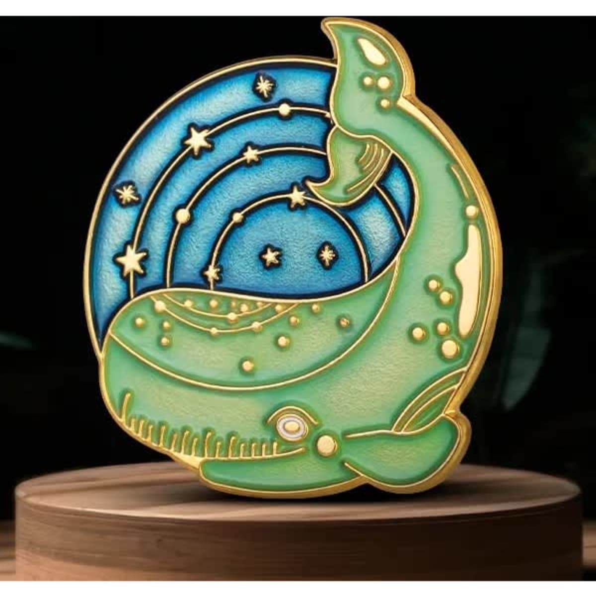 A lapel pin featuring a whale design in shades of green and blue, set against a starry background.