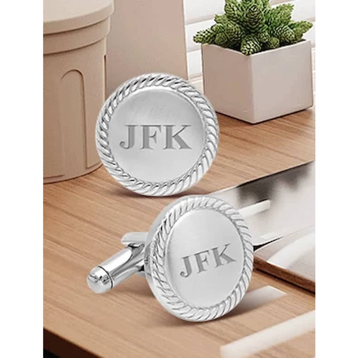 Custom engraved silver cufflinks with a rope design and initials, placed on a wooden surface with potted plants.