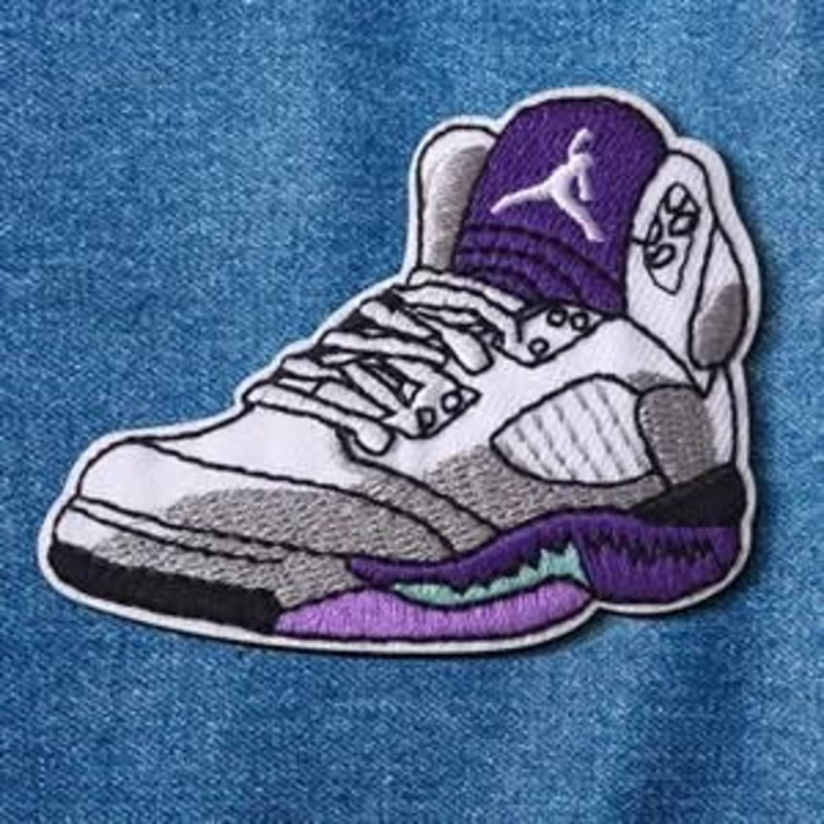 An embroidered patch of a sneaker, featuring purple, white, and grey colours, with a logo.