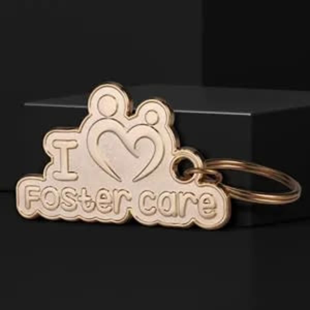 A gold 2D die struck key chain featuring the text "I ❤️ Foster care" and a heart design, on a dark background.