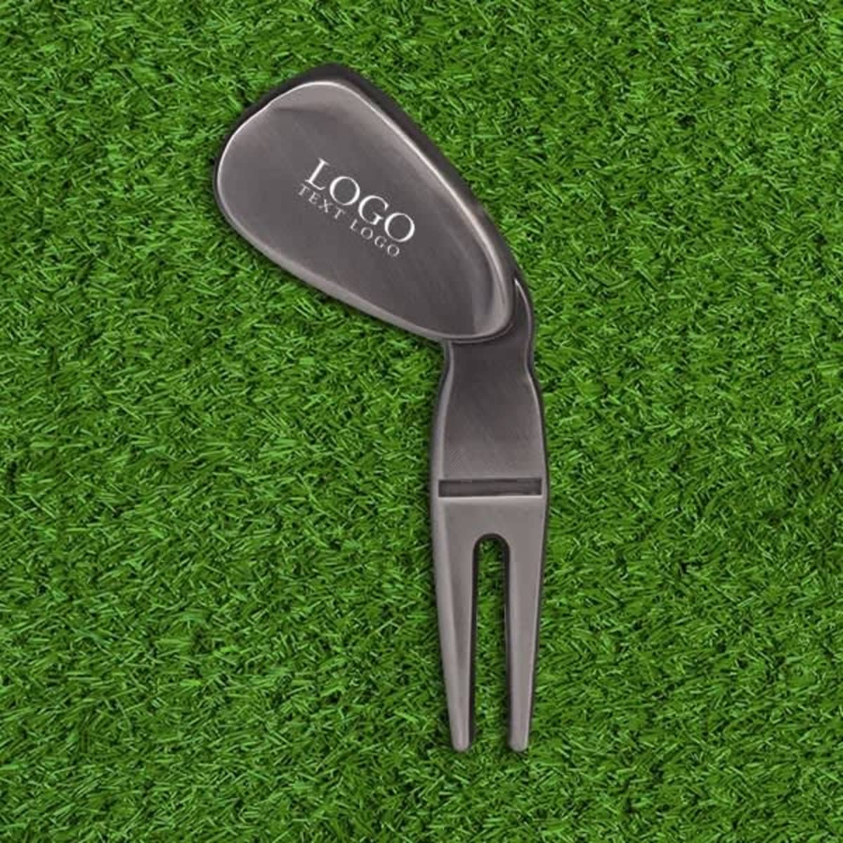 A black divot tool with a forked end, placed on green grass, featuring a logo.