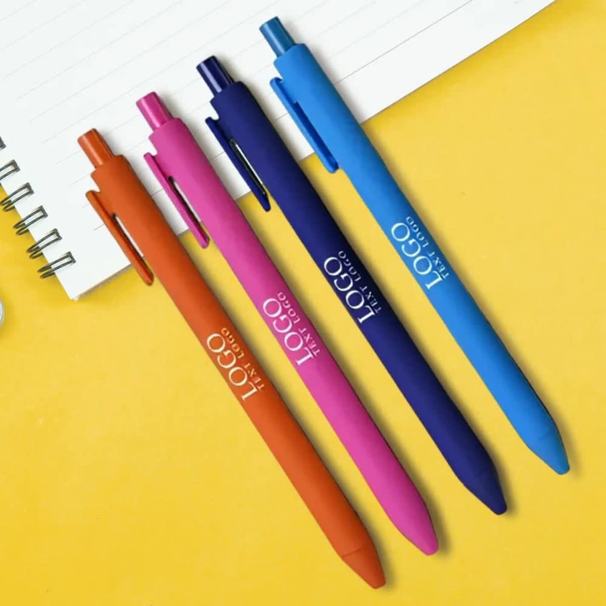 Four gel ink pens in vibrant colours: orange, pink, blue, and purple, positioned next to a notebook. The pens have branding.