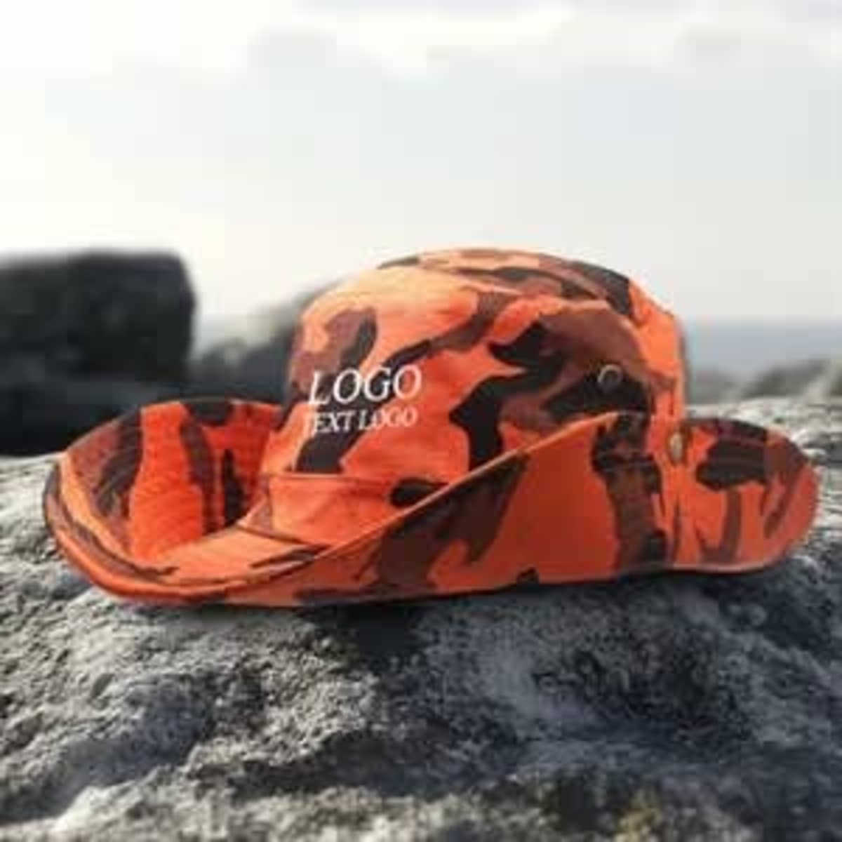 A large brim bucket hat in orange camouflage with a logo, placed on a textured rock surface.