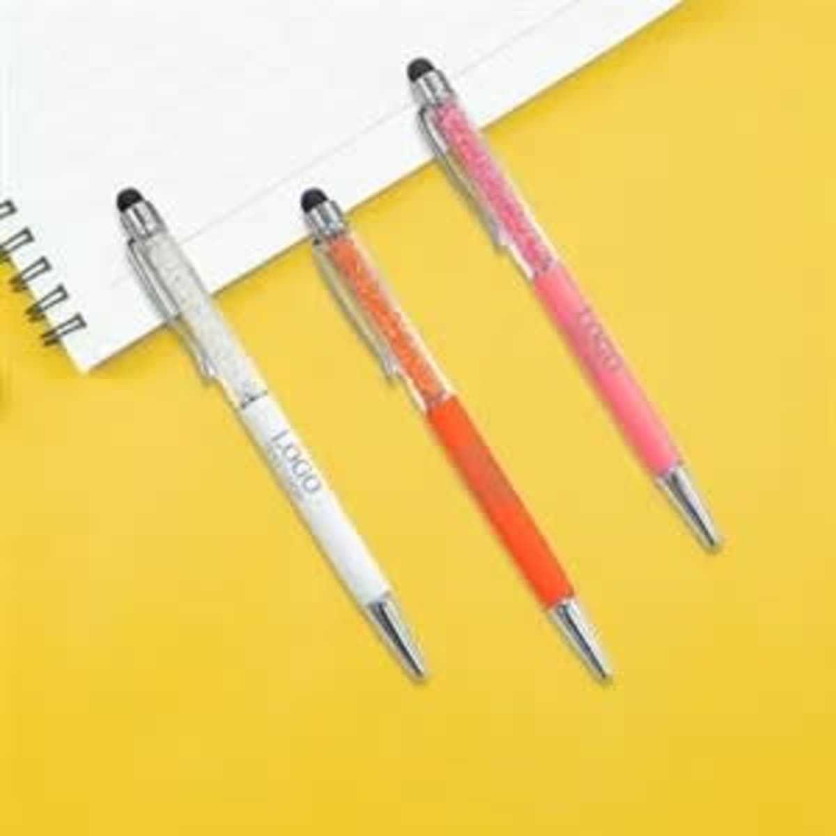 Three retractable ballpoint pens in white, orange, and pink, featuring stylus tips, placed beside a notepad.