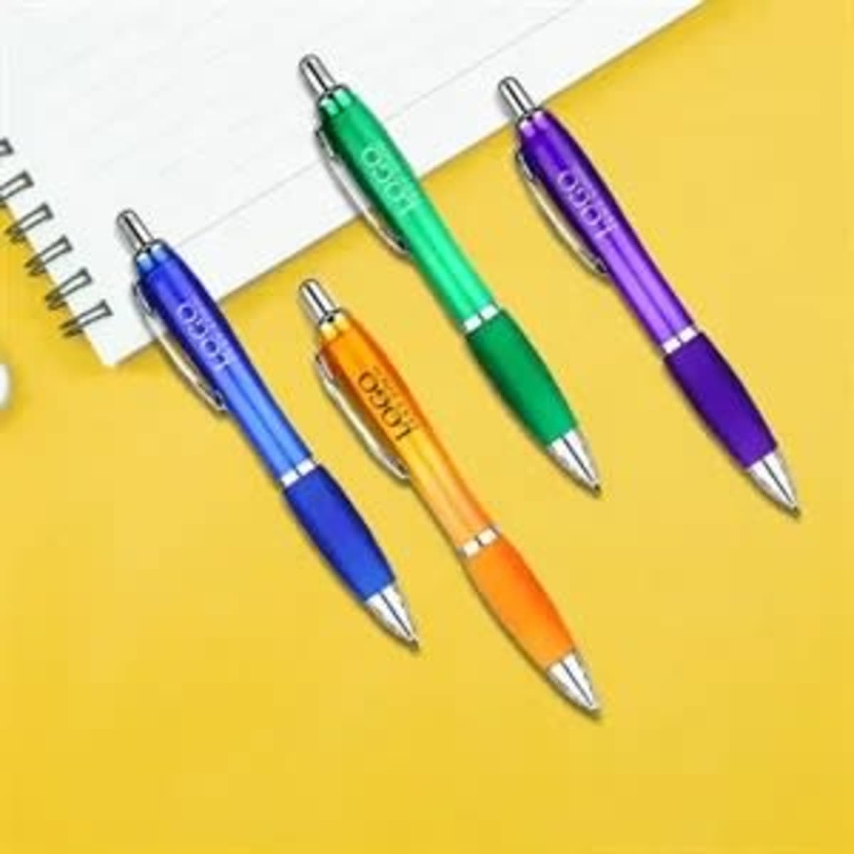 Four plastic satin pens in blue, green, orange, and purple, displayed on a yellow background next to a notepad.