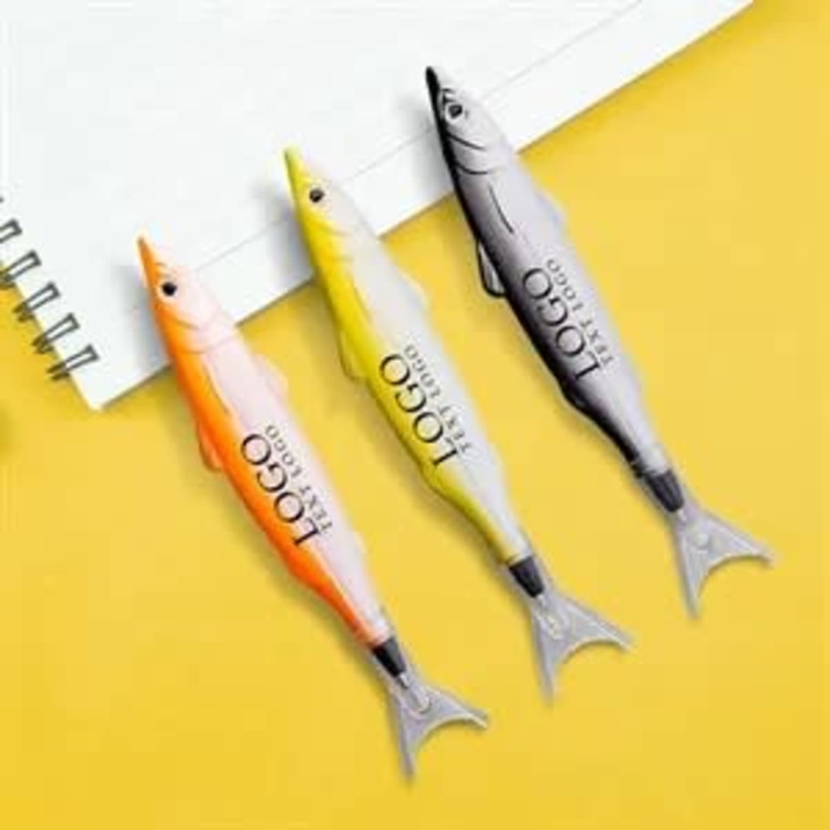 Three fish-shaped pens in orange, yellow, and grey, placed on a yellow background beside a notepad. Each pen has a logo.