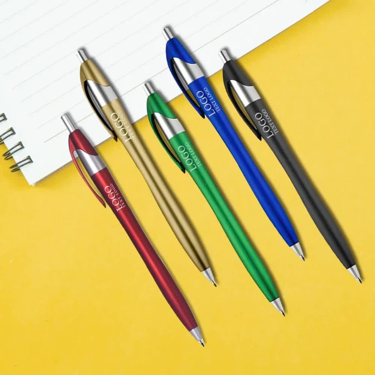 A set of five plastic ballpoint click pens in red, blue, green, gold, and black with metallic accents beside a notepad.