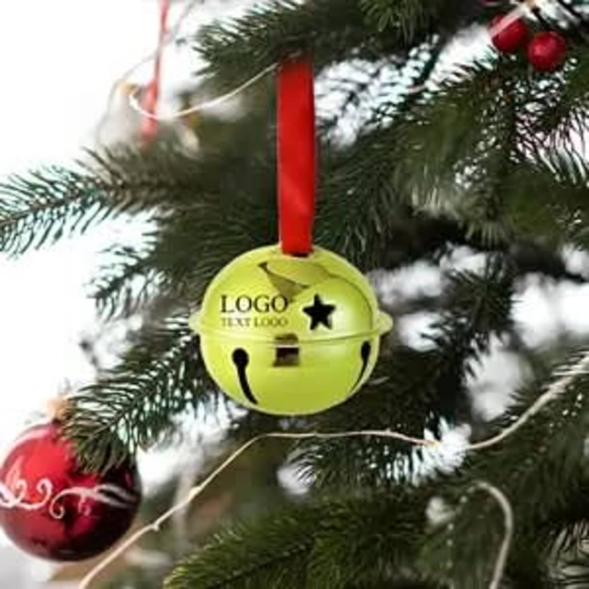 A green jingle bell ornament with a red ribbon, hanging on a Christmas tree branch, alongside red ornaments.