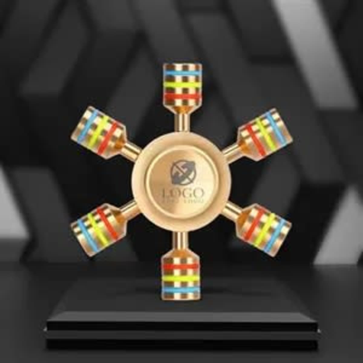A gold hexagonal fidget spinner with colourful detachable sections, displayed on a black pedestal with a logo.
