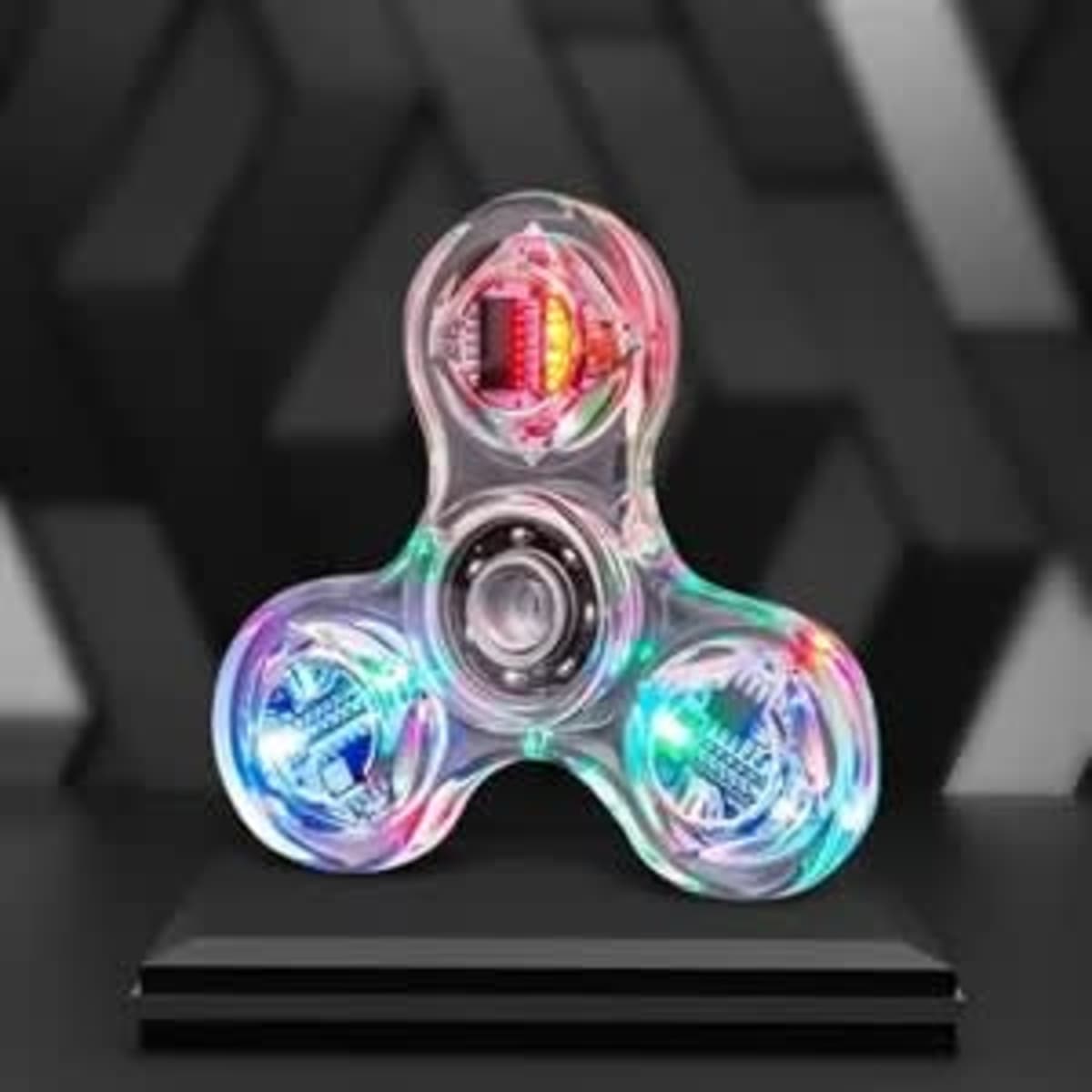 A clear LED light fidget spinner with multicoloured lights against a dark geometric background, displayed on a stand.