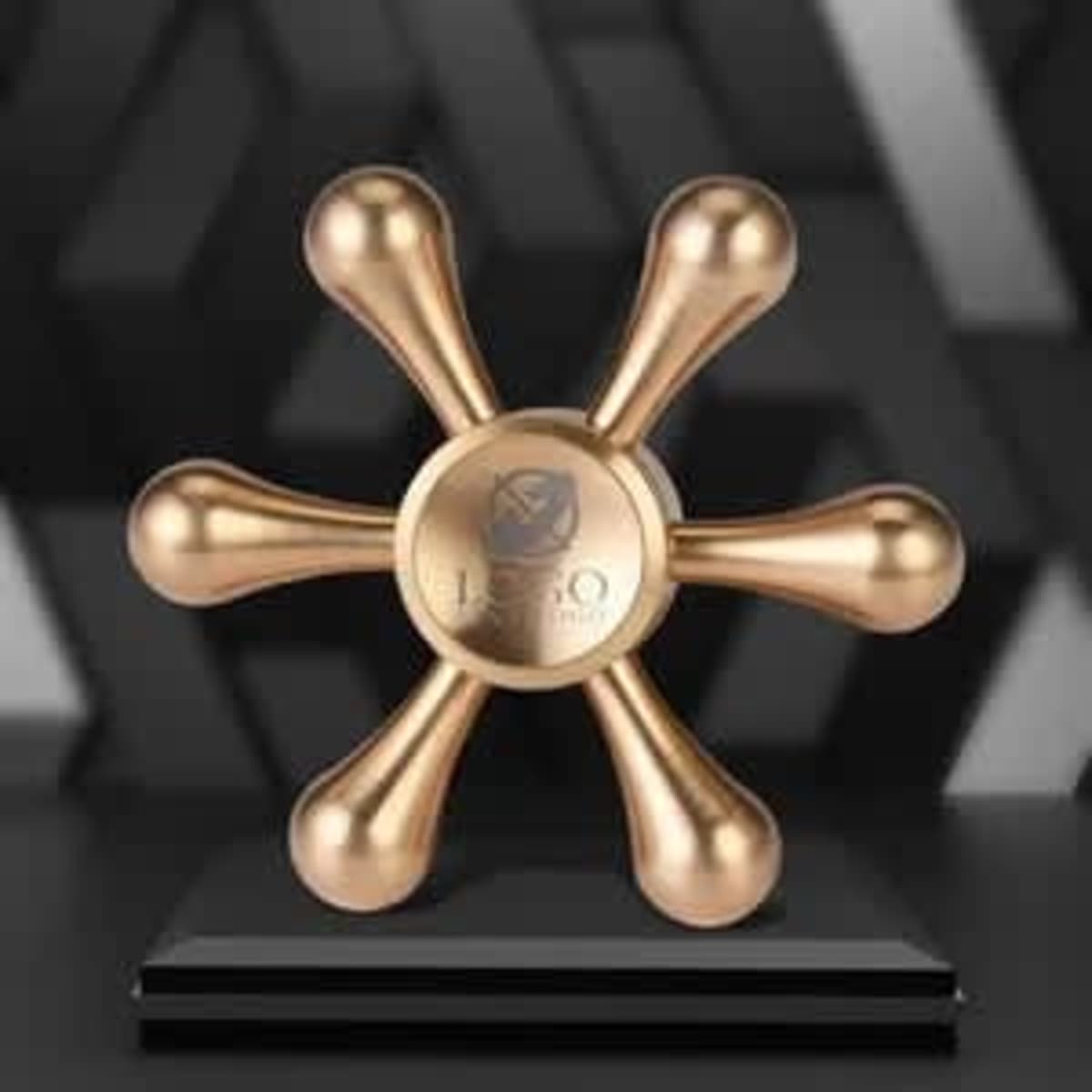 A bronze fidget spinner in a playful rudder shape, displayed on a black pedestal. Features a logo at the centre.