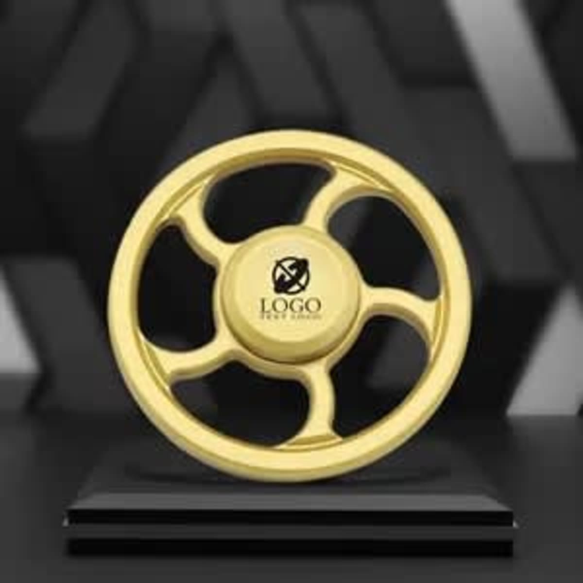 A gold metal fidget spinner with a central logo, displayed on a black pedestal against a textured background.