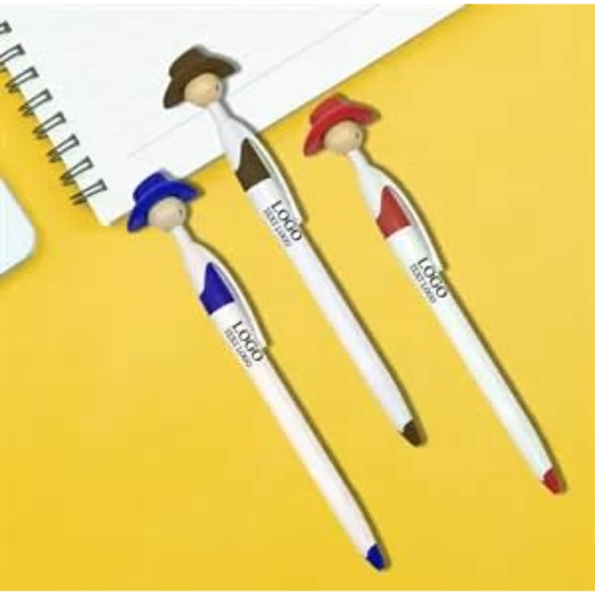 A set of three pens with cowboy figure tops in blue, red, and brown, resting on a yellow background with a notepad.