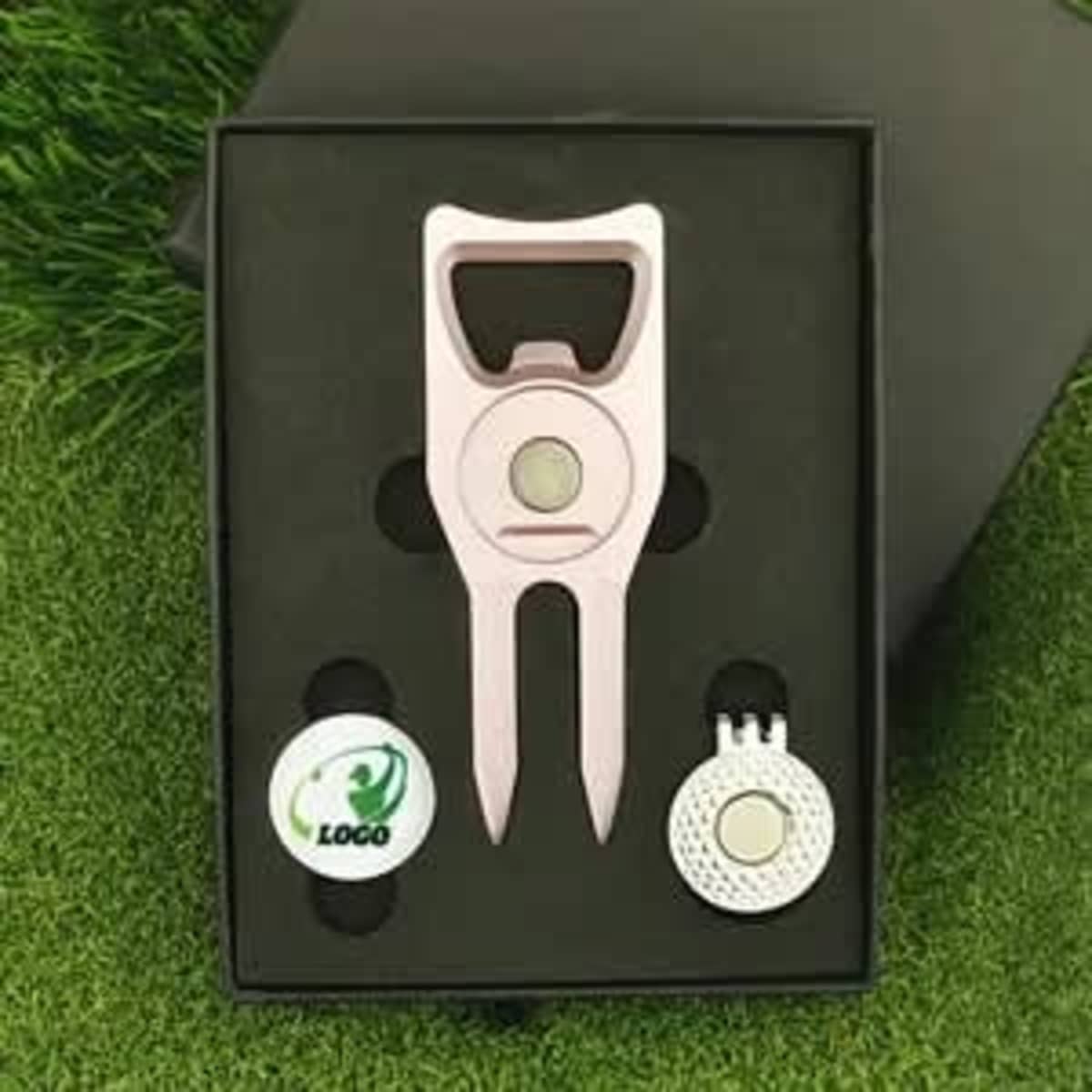 A gift box set featuring a pink cap clip, a ball marker, and a circular marker, presented on a grass background.