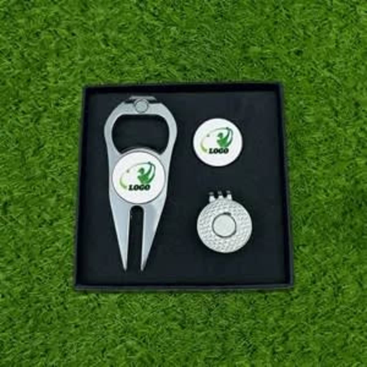 A silver golf tee marker, a ball position marker, and a clip presented in a black box on grass.