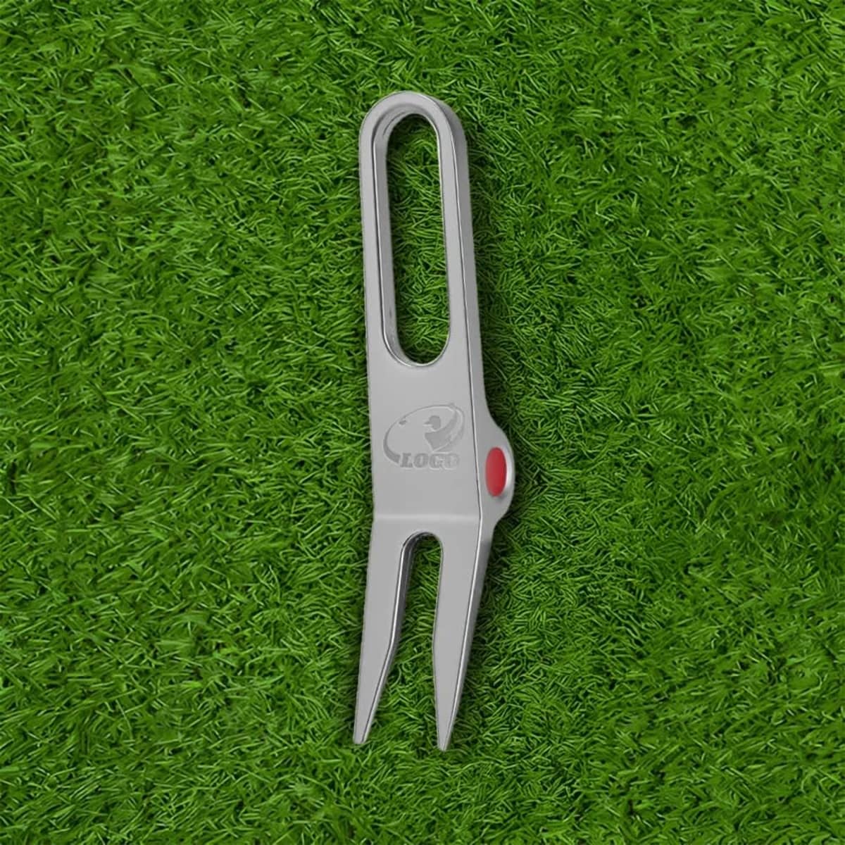 A metal golf divot tool in grey with a red accent, resting on green grass. The tool features a logo.