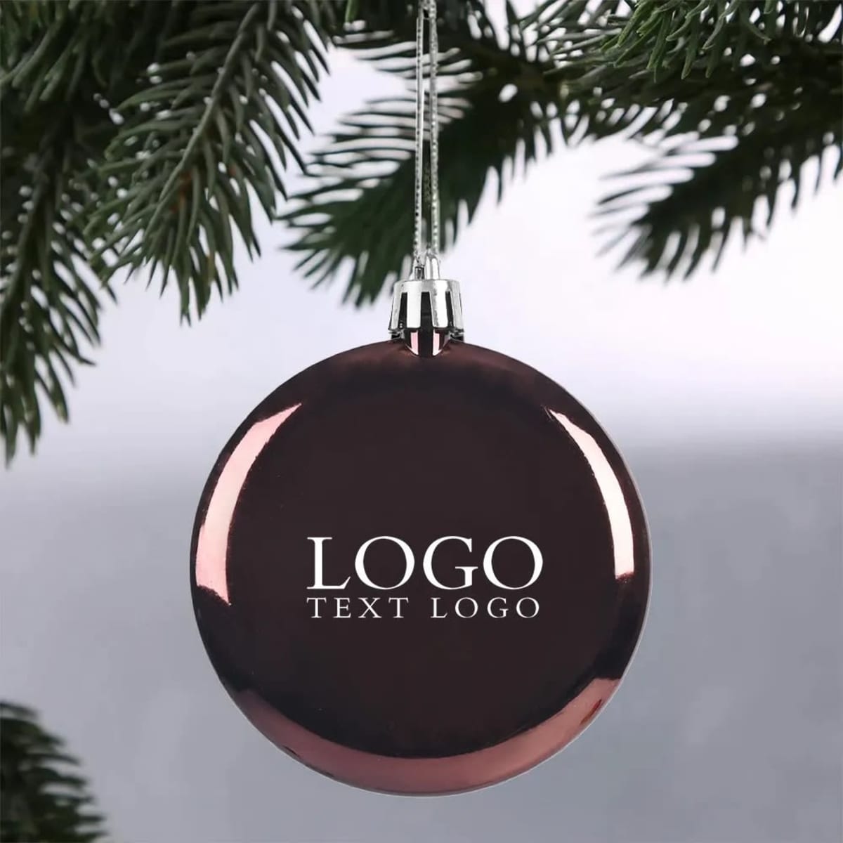 A shatter-resistant flat round Christmas ornament in a dark colour, featuring a logo, hanging from a tree branch.