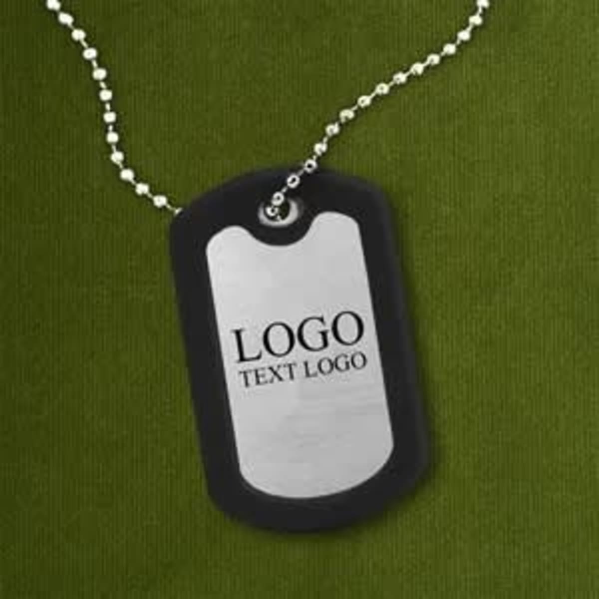 A square metal dog tag with a silicone edge, featuring a logo, attached to a beaded chain against a green background.
