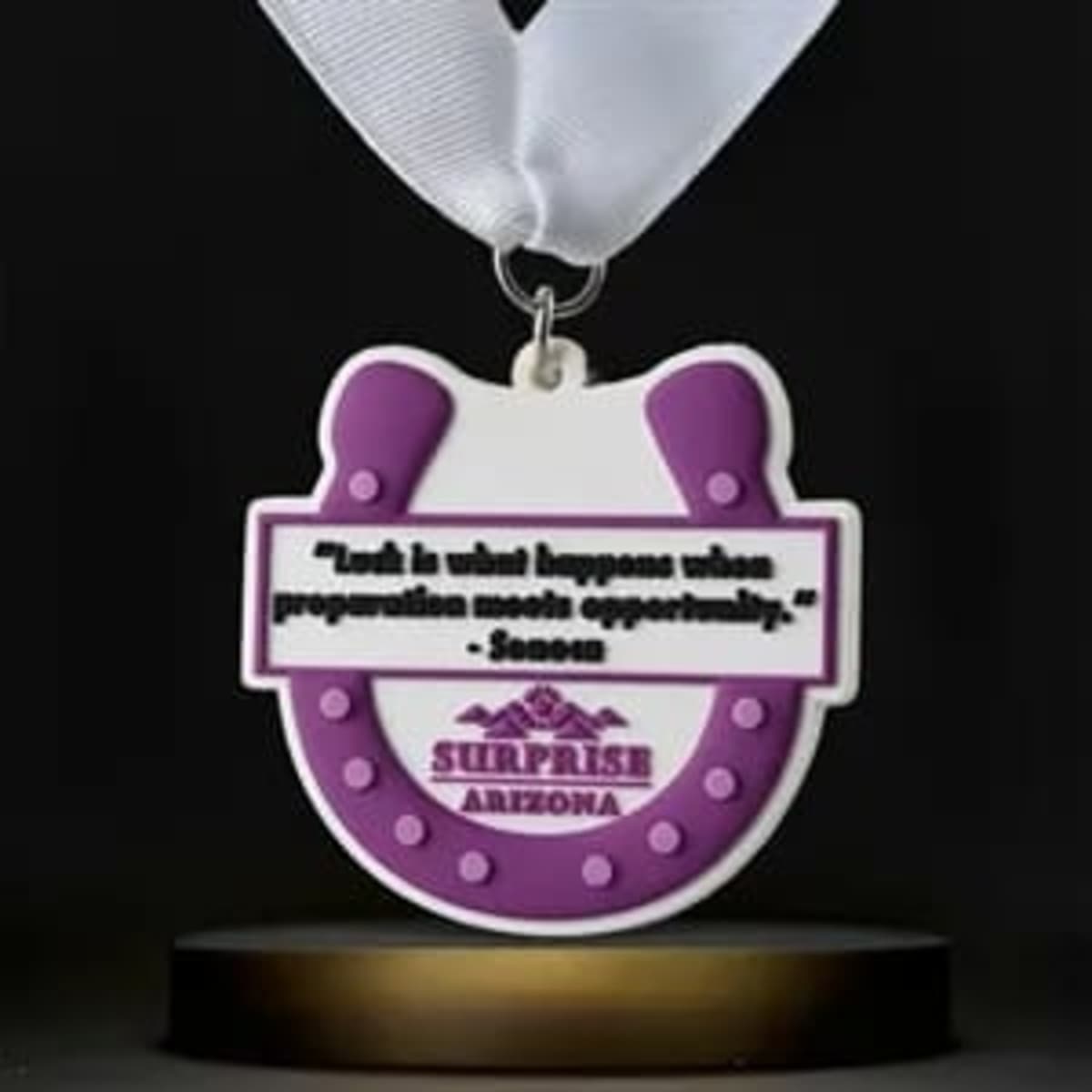 A round purple and white soft PVC medal with a ribbon, featuring a horseshoe design and a quote.