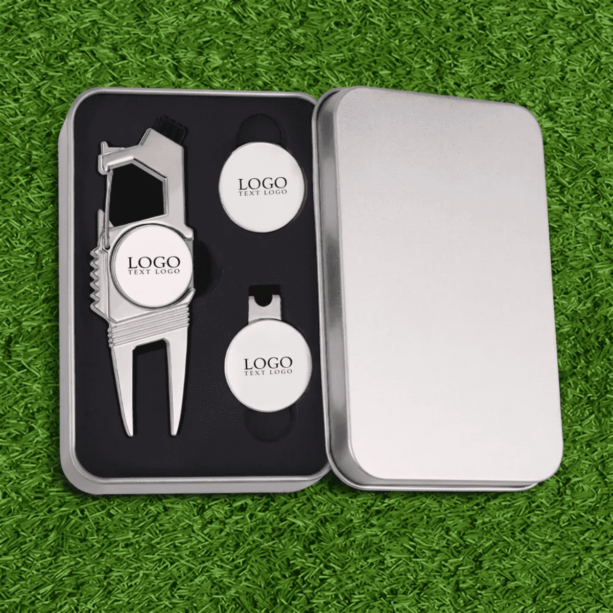 A silver gift box containing a divot tool and two ball markers, all with a logo, set against artificial grass.