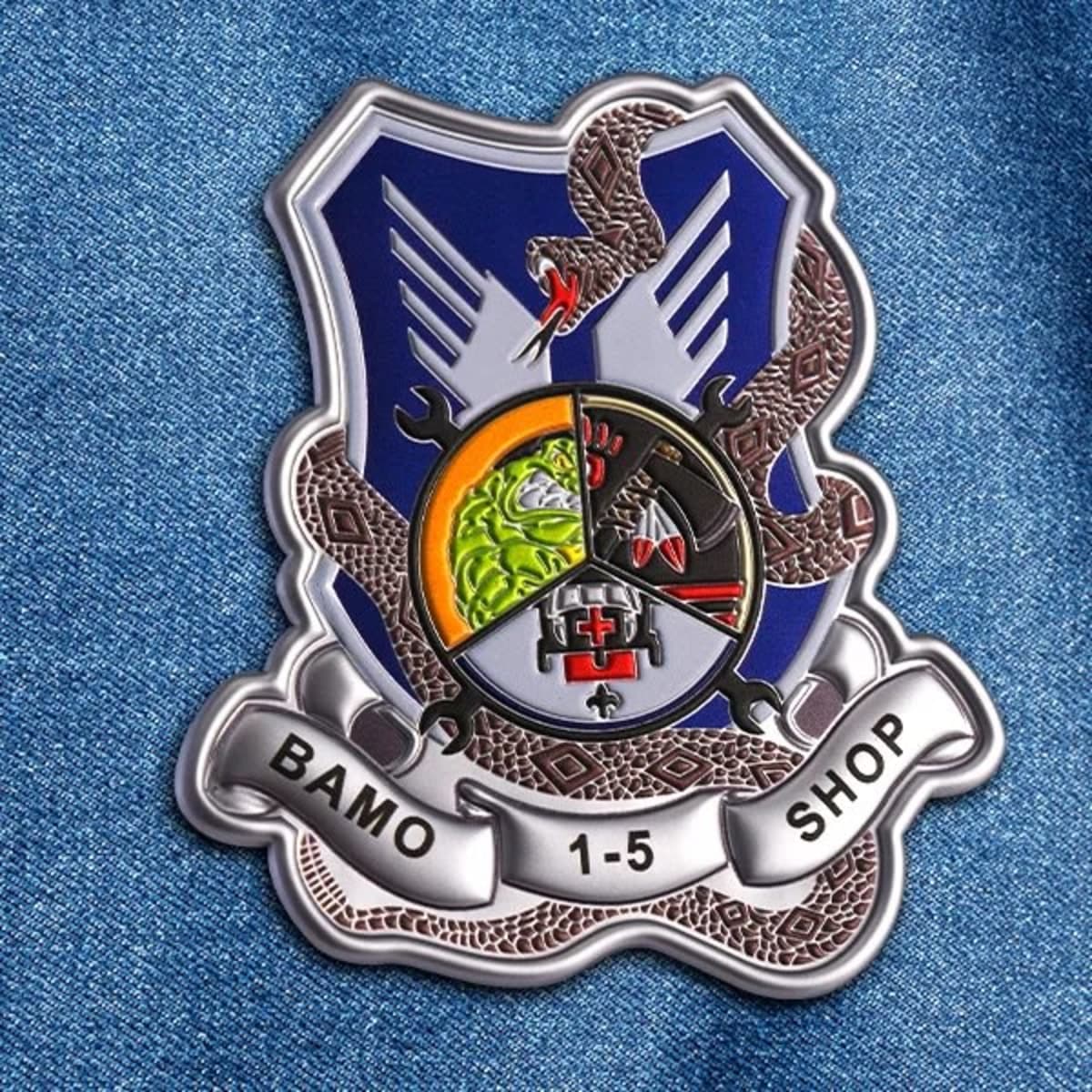 A metallic badge featuring a blue shield, intricate designs, and a logo. It has a snake and banner at the base.