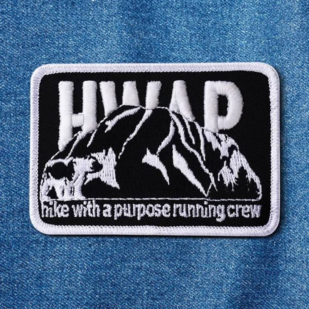 A black and white embroidered patch featuring a mountain design and the text "HWAP". The patch has a white border.