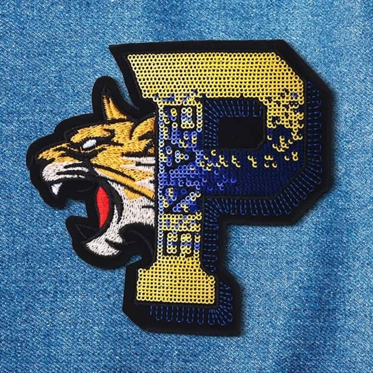 A sequin patch featuring a yellow and blue 'P' with a tiger's head design, set against a blue denim background.
