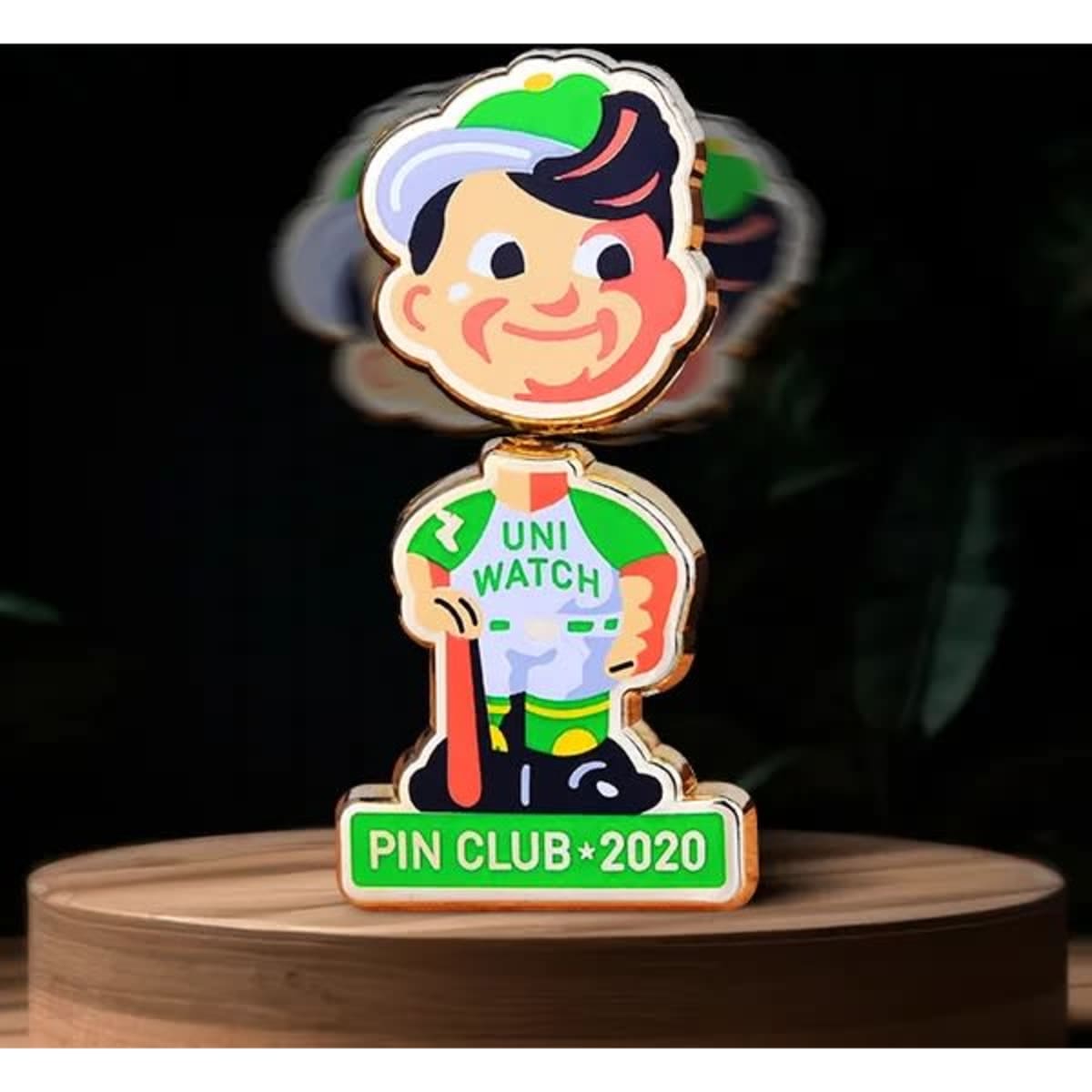 A colourful bobblehead pin featuring a cheerful character in green and white attire, with a branded logo at the base.