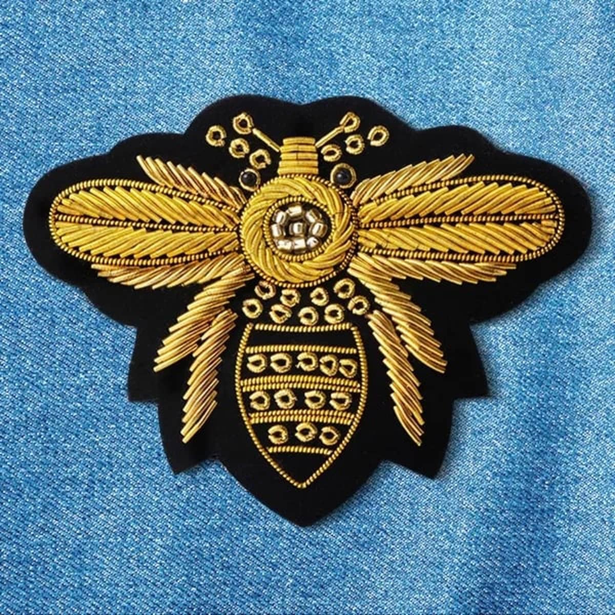 A gold and black embroidered patch in the shape of a bee, set against a blue denim background.