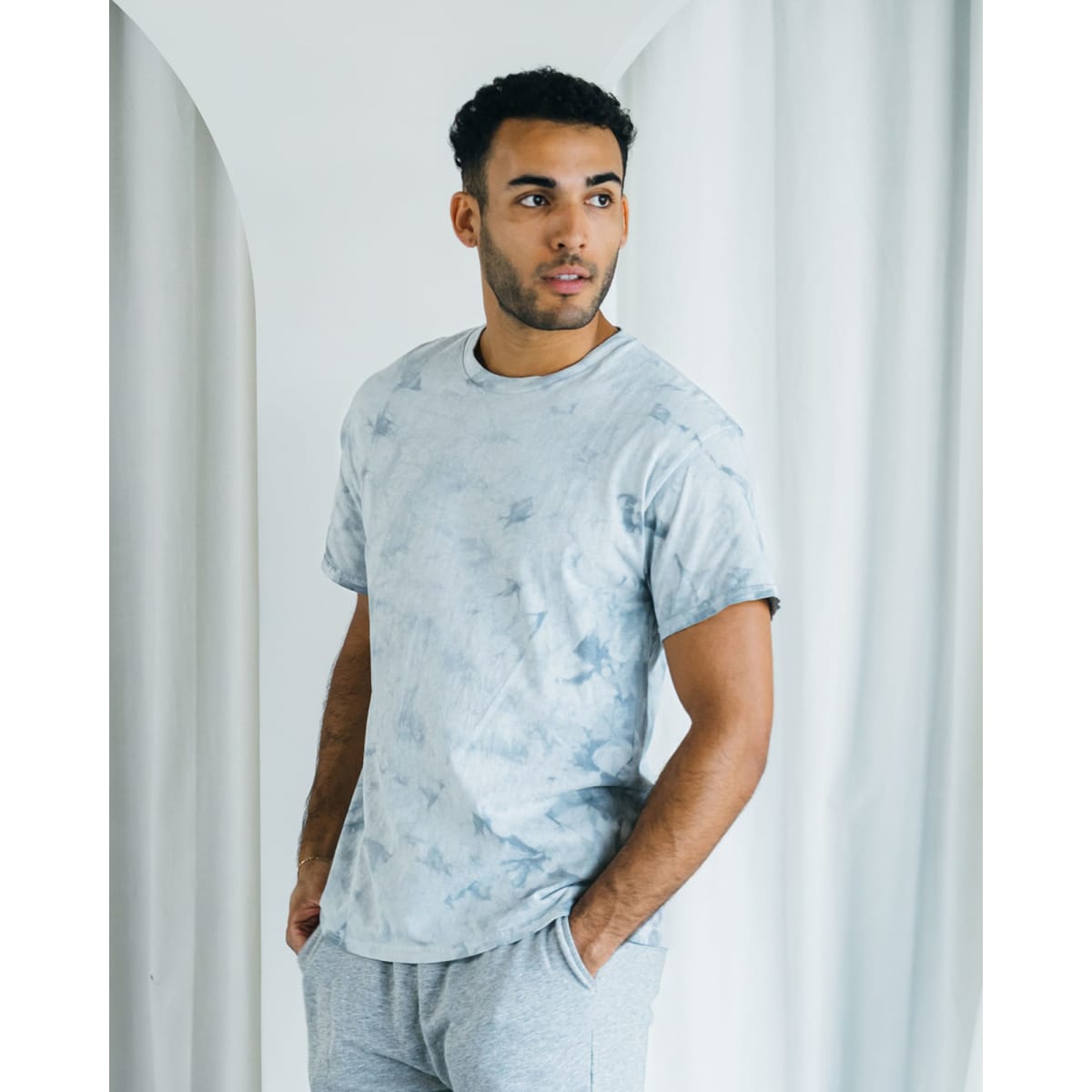 A man wearing a grey tie-dye t-shirt and light grey shorts, standing against a soft, neutral background.