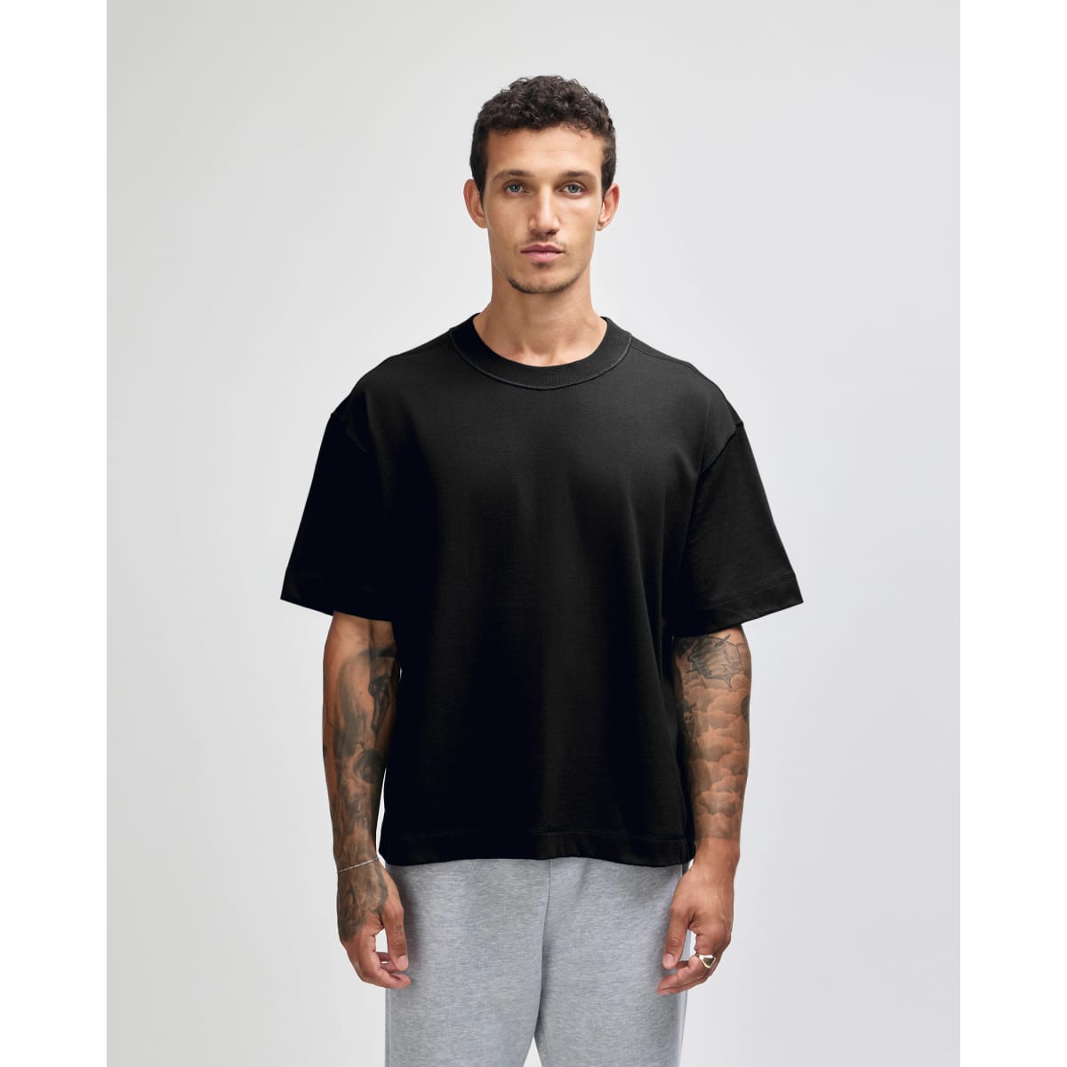 A black unisex heavyweight cotton boxy t-shirt displayed on a model with tattoos, paired with grey trousers.