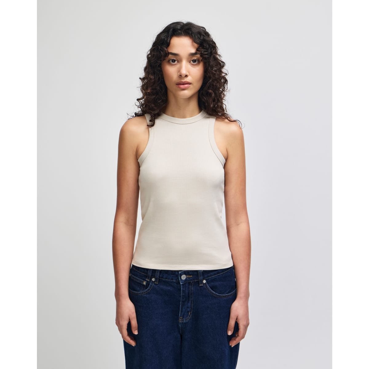 A fitted tank top in a light beige colour, featuring a high neckline and without sleeves. The model is wearing jeans.